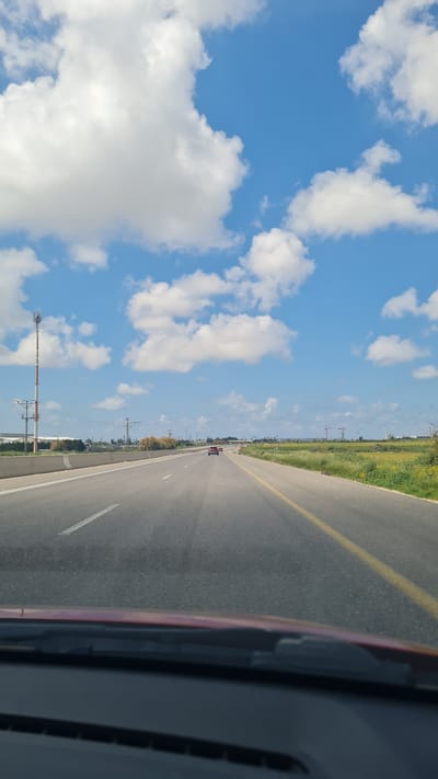 The image depicts a road, likely within or bordering the Hof Ashkelon Regional Council, Israel, as inferred from the surroundings. The road stretches out toward the horizon, with a yellow line marking the shoulder and a dashed white line separating lanes. A car is visible ahead on the road. Vegetation borders the road on one side, suggesting an agricultural or rural area. A communication tower stands nearby. The sky is blue with puffy white clouds, indicating a clear day. The image is taken from within a vehicle, with part of the car's interior visible.