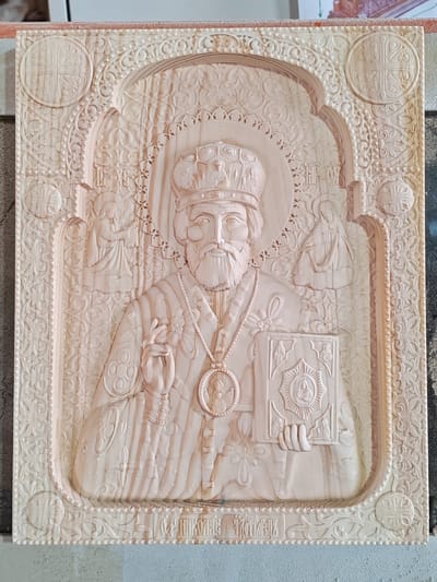This is a detailed photograph of a carved wooden icon. The icon depicts a central figure, likely a saint or religious leader, wearing a crown and ornate robes, holding a book in one hand and making a gesture with the other. The central figure is surrounded by intricate carvings, including decorative borders, smaller figures within arched niches, and religious symbols such as crosses. The wood appears to be light-colored, possibly pine or linden, and exhibits a fine grain. The carving is deeply incised, creating a strong sense of relief. At the bottom of the icon, there is an inscription in Cyrillic script. The photograph is taken from a slightly elevated angle, showing the texture and depth of the carving. The background is out of focus, with hints of what might be a workshop environment. The overall impression is of a handcrafted, religious artwork. This icon is located in the city of Ичетовкины, Russia.