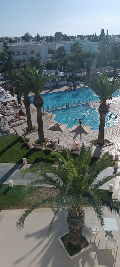 This is a high-angle, sunny daytime outdoor photograph of a swimming pool area at a resort in Porto El Kantaoui, Tunisia. The pool, filled with blue water, is in the center of the frame, with several people swimming and playing. Palm trees are prominently featured throughout the scene, with some casting long shadows on the tiled patio. Around the pool are lounge chairs, some with umbrellas. In the background, white buildings with flat roofs, characteristic of the architecture in the region, are visible, along with lush green trees. The overall atmosphere suggests a vacation resort setting.
