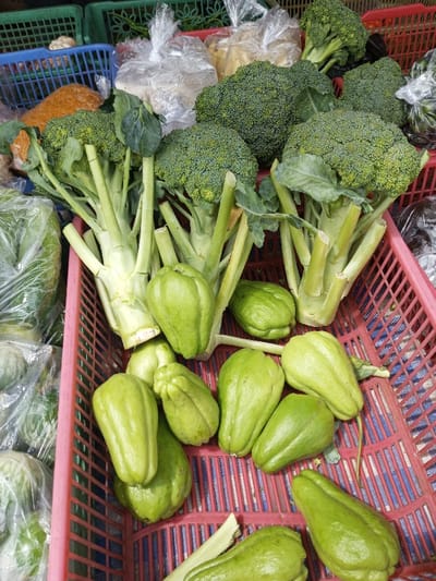 The image displays a close-up view of fresh produce arranged in plastic woven baskets, located at a market in Taohom, Laos. The primary subjects are multiple heads of green broccoli with dense florets and thick, pale green stalks, predominantly in the upper and mid-sections of the frame. Below the broccoli, numerous light green, pear-shaped chayote squash are visible, primarily in a pink plastic basket. In the background, additional green and blue plastic baskets are present, along with several clear plastic bags containing various other food items, including one with an orange-brown granular substance and others with lighter-colored contents, suggesting a diverse market stall. No people, actions, or direct interactions are depicted. The scene is well-lit, highlighting the textures and colors of the vegetables.