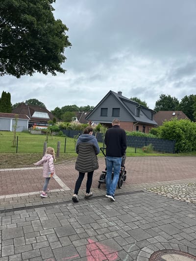 This image depicts a family of two adults and a young child walking along a paved sidewalk in a residential area of Deinste, Germany, under an overcast sky.

The adult male, viewed from behind, wears a dark jacket and blue jeans, pushing a black stroller with white-rimmed wheels. To his left, an adult female, also seen from behind, wears a dark olive-green quilted jacket with a grey hooded interior and dark trousers. A young blonde girl, wearing a light pink patterned jacket, grey pants, and pink athletic shoes, walks slightly ahead and to the left of the adults, holding a small, colorful object in her right hand.

The ground consists of a pedestrian pathway segmented into two types of pavers: a reddish-brown section closer to the street and a grey section closer to the properties. A metal drainage grate runs between these sections. Red and white chalk markings are present on the grey pavers, and a metal manhole cover is partially visible in the lower right corner.

A dark grey horizontal slatted fence separates the sidewalk from the residential properties. Behind the fence, a prominent two-story house on the right features a dark grey roof and upper exterior walls with dark window shutters, contrasting with a brick lower story. Further to the left, a house with a brown tiled roof and a white dormer window is visible, along with a light grey shed or garage. Mature green trees and dense bushes are scattered throughout the background, partially obscuring the houses and creating a verdant environment. The sky is uniformly grey and cloudy.