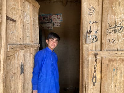 The image shows a young man standing in the doorway of a building. He is wearing a blue shirt and is looking directly at the camera. The doorway is made of wood and has a padlock on it. The building is located in Saranan, Pakistan, at the coordinates 30.532, 66.909. The walls of the building are made of mud brick. The door has numerous Arabic inscriptions on it.  There are a number of posters on the wall behind the young man, with pictures of unidentified people.  There is a chain attached to the padlock hanging on the door.  The environment is dusty and barren.