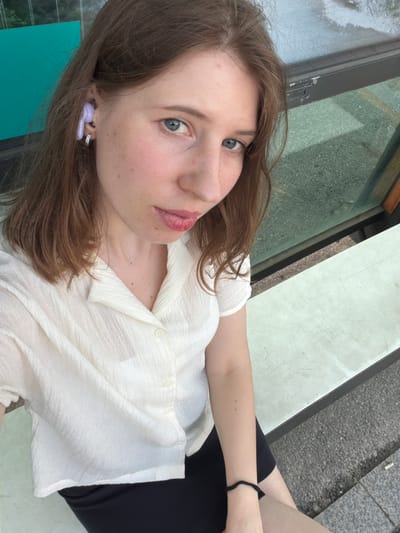 The image shows a young woman with brown hair and blue eyes, wearing a white collared shirt and a black skirt. She is sitting on a bench, possibly at a bus stop, with a teal-colored wall or structure behind her and a glass partition to her right. She has a purple earbud in her left ear and a silver hoop earring. The scene appears to be outdoors, with the ground visible in the foreground showing gravel and concrete. The lighting suggests it is daytime. The overall impression is of a casual, everyday moment captured in an urban or semi-urban environment.