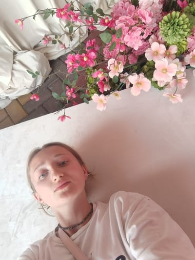 The image is a selfie taken from a high angle, featuring a young woman lying on a white surface, likely a bed or couch. She has fair skin, light-colored hair, and is wearing a white t-shirt and a dark beaded necklace. Her expression is neutral, and she is looking directly at the camera.

To her upper right, there is a vibrant floral arrangement with various shades of pink and green flowers, some of which appear to be artificial. The flowers are on branches with green leaves.

In the background, to the upper left, a draped, light-colored fabric creates a soft, blurred backdrop. Below the flowers and to the left, there is a section of paving stones, indicating an outdoor or semi-outdoor setting.

Based on the provided image, there is no information that would allow for the determination of the specific location in the city of Starobzhegokay, Russia.