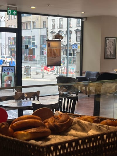 The image presents an interior view from a Caffe Nero establishment, looking outwards through a large glass window onto a city street in Belfast, United Kingdom, specifically identifiable near Alfred Street.
In the immediate foreground, a dark brown wicker basket contains an assortment of baked goods, including several golden-brown oblong pastries and a darker, braided pastry, arranged on a white patterned paper liner. A portion of a red circular object is visible on the far left edge.
Beyond the foreground counter, interior elements of the cafe include dark wooden tables with circular tops and accompanying dark wooden chairs. Further inside, a dark grey upholstered armchair and a dark patterned armchair are visible. A framed rectangular artwork, depicting a stark building silhouette with a single figure, hangs on the right-hand wall. A Caffe Nero advertisement board for "ICED HONEY FOAMED AMERICANO" is positioned near the window on the left. A banner advertising "SEASONAL COFFEE BRAZIL SINGLE ORIGIN" hangs suspended from the ceiling by chains. An illuminated green "EXIT" sign is affixed above the window frame on the upper left. Reflections of internal cafe signage, including words like "MOCHA," "LATTE," "CAPPUCCINO," and "PASTA," are visible on the window glass.
Through the window, a city street is observed with parked or idling vehicles, including a white SUV on the left. Two individuals in light blue tops are visible near the SUV. Red and white temporary barriers and other equipment indicate ongoing roadworks. Various commercial establishments line the street, notably multiple "Greggs" signs and a building sign for "THE VANTAGE." A prominent blue street sign post points towards "ALFRED ST PUBLIC" and "ASSEMBLY ROOMS." The background features multi-story urban buildings under daylight conditions.