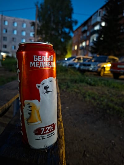 A close-up, low-angle shot of a red and white beer can resting on a dark, textured surface, possibly a wooden railing. The can is oriented horizontally, with the label facing the camera. The label features a cartoon illustration of a white bear holding a frothy mug of beer. Prominently displayed on the can is the text "БЕЛЫЙ МЕДВЕД" (White Bear) in large, bold, white letters. To the right of the bear illustration, a red brushstroke highlights the text "7.2%", indicating the alcohol by volume. Below this, smaller text in Russian reads "КУЛЬТ МЕДВЕД" (Cult Bear) and "ЖИГНИ ДЕНЬ" (Live Day).
The background is blurred, suggesting a shallow depth of field. On the left, there's a hint of a building with illuminated windows, set against a dark blue twilight sky. To the right of the building, the background transitions to dark, textured ground with some green foliage, possibly grass or bushes. The overall lighting suggests it is dusk or evening. The image's perspective and the blurred background create a sense of intimacy with the beer can, while the ambient light hints at an outdoor setting in an urban or semi-urban environment. No people are visible. The scene evokes a casual, possibly relaxing mood. The location context provided is Ridder, Kazakhstan.