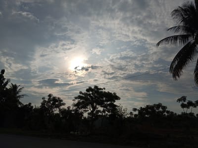 A view of the sky over Cotpuuk, Indonesia, features a bright sun peeking through scattered clouds. The lower portion of the image shows silhouettes of trees and foliage against the sky, with a power line stretching horizontally across the frame. A paved road is visible in the foreground. The scene appears to be captured during daytime, possibly early morning or late afternoon due to the low angle of the sun and the overall lighting.
