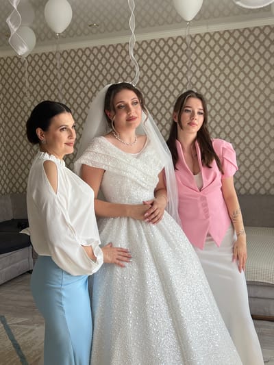 Three women stand in a room with patterned wallpaper. The woman on the left wears a white blouse with cut-out shoulders and a light blue skirt. The woman in the center is a bride in a white sequined wedding dress with a veil and pearl necklace. The woman on the right wears a pink blazer over a white top and a white skirt. She has a tattoo on her arm. White balloons are visible in the upper part of the frame. Furniture including sofas and a rug are present in the background. The scene appears to be indoors, likely in a home or event space, within the city of Astrakhanka, Kazakhstan.