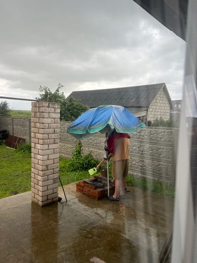 In Бучацінскі сельскі Савет, Belarus, on a rainy day, two individuals stand under a blue and green umbrella near a brick barbecue. One person, wearing a red garment over a white shirt and black pants, holds a green watering can, appearing to pour water onto the barbecue coals. The other individual, dressed in a tan shirt and beige shorts, stands beside them. The ground is wet, reflecting the overcast sky. A partially constructed brick pillar stands to the left, and a concrete fence forms the background. Behind the fence, a house with a grey roof and a grassy area are visible.
