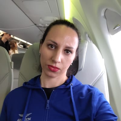 The image depicts a young adult female taking a selfie inside an airplane cabin, located in Skulte, Latvia. The subject is positioned centrally, looking directly at the camera, with a neutral expression and slightly pursed lips. She has dark hair pulled back, prominent eyebrows, brown eyes with eyeliner, a small stud piercing on her right nostril, and is wearing red lipstick. She is dressed in a royal blue hooded zip-up sweatshirt with white embroidery on the left chest, partially visible as "AVI OM" and a stylized wing or bird design. Small silver-colored stud earrings are visible in both ears.

The background shows the interior of a commercial aircraft cabin. To the left of the subject, another passenger, a young male wearing a light-colored baseball cap and a dark shirt, is visible facing away. Further back, a female with light brown hair is seated. The cabin features light-colored, possibly off-white or light grey, seats with quilted stitching patterns. White overhead panels line the ceiling, with linear green accent lighting strips running along the upper cabin walls. Bright natural light enters the cabin from a window visible on the right side of the frame.