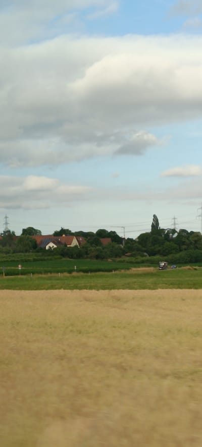 A horizontal, eye-level shot captures a bucolic landscape in Schwaikheim, Germany. The foreground is dominated by a field of dry, golden grass, blurred by motion, suggesting a moving viewpoint. Beyond this, a strip of vibrant green field extends towards a treeline. In the mid-ground, nestled amongst lush green trees, are several houses with terracotta-tiled roofs and white walls, indicating a rural or semi-rural settlement. A few solar panels are visible on one of the rooftops. Power lines and transmission towers punctuate the horizon behind the houses, hinting at the proximity of infrastructure. Two vehicles, a dark truck and a blue car, are visible on a road that runs parallel to the treeline. The sky above is a mix of pale blue and scattered white clouds, with larger, more diffuse clouds in the upper portion of the frame. The overall impression is one of a tranquil, sunlit day in the German countryside.