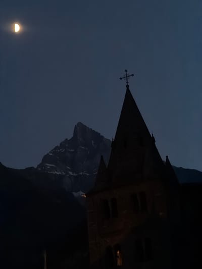 This media file depicts a dramatic nighttime scene featuring a gothic-style building perched precariously on a cliffside, with a crescent moon low in the sky. The building, possibly a church or a castle, is dark and imposing, with its sharp angles and steep roofline silhouetted against the twilight sky. A cross adorns the peak of one of its spires. The cliff face is rugged and steep, suggesting a mountainous or high-altitude location. The overall mood is one of mystery and awe, amplified by the darkness and the dramatic setting. The faint glow of lights within the building hints at occupancy, contrasting with the surrounding emptiness. The image appears to be captured during either dusk or dawn, given the presence of the moon and the dim lighting. The location context of Saint-Maurice, Switzerland, adds a layer of historical and geographical specificity to the scene, evoking images of the Swiss Alps and their ancient structures. No visible text is present in the image.