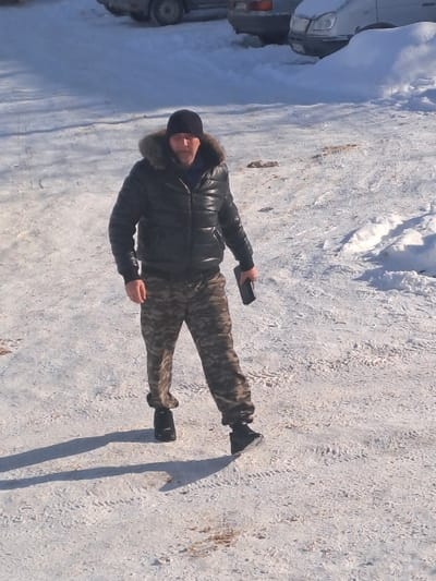 The scene, located in Bolshaya Elhovka, Russia, depicts an adult male walking on a snow-covered, cleared outdoor surface under bright sunlight. The man, positioned centrally, wears a black puffer jacket with a fur-lined hood, a black beanie, camouflage-patterned trousers, and dark boots. He holds a dark rectangular electronic device, consistent with a smartphone, in his right hand at waist level. Distinct tire tracks are visible on the icy and snow-covered ground. In the background, partially obscured by snow, are two parked vehicles: a light-colored sedan or hatchback and a darker vehicle. Long shadows are cast to the left of the subject, indicating a low sun angle. No other individuals or interactive subjects are present.