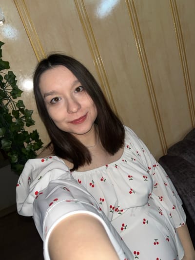 An indoor scene in Kozlovka, Russia, captures a young adult female in a selfie. She possesses shoulder-length dark brown hair, brown eyes, and fair skin, and is seen smiling faintly directly at the camera. She wears a white, off-the-shoulder top or dress adorned with a repetitive red cherry pattern, complemented by a thin, silver-colored chain necklace. Her right arm is extended, holding the camera. The background consists of a light tan wall with vertical decorative paneling that exhibits a glossy sheen under artificial lighting. An artificial green leafy vine is visible on the left side of the frame. A portion of a dark-colored, upholstered piece of furniture, likely a couch, is visible on the right. The setting is consistent with a private residence.
