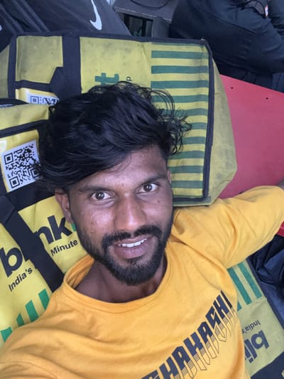 A dark-skinned man with a beard and dark hair is depicted from a slightly elevated angle, lying on his back amidst several yellow and black delivery bags in Palghar, India. He wears a bright yellow short-sleeved t-shirt with dark, stylized text visible on the chest, partially readable as "BHARATHI" or similar. The man is smiling, showing his upper teeth, and looking directly at the camera.
The delivery bags are yellow with black accents. On the left side of the frame, one bag prominently features the text "blnk" and "India's Minute," indicative of the Blinkit quick commerce service. A white sticker with a black QR code is affixed to this bag. Another delivery bag, visible on the right, displays green horizontal stripes against a yellow background. In the upper left background, a black bag with a white Nike swoosh logo is partially visible. A bright red surface is present on the right side of the frame, adjacent to the striped bag and the man's arm.