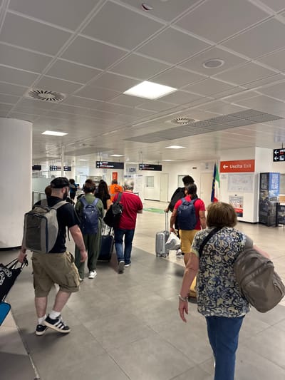 A group of people are queued with luggage at an airport in Cinisi, Italy. They are lined up on a gray floor beneath a white tile ceiling. A sign indicates "Uscita Exit". There is an Italian flag hanging.