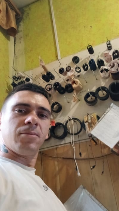 The media file features a close-up, tilted selfie of a person with short, dark hair and a fair complexion. They are wearing a white t-shirt. A tattoo is visible on the person's neck. The background is a cluttered surface displaying numerous small, black, circular objects, many of which are mounted on pins. These objects appear to be various types of electrical components, possibly inductors or resistors, arranged on what seems to be a workbench or a display board. Some of these components are partially wrapped in fabric or paper. White wires and cables are also present, some connected to the components and others trailing across the scene. To the left of the frame, a wooden surface is visible, possibly a table or wall paneling. The overall setting suggests a workshop or storage area. The lighting is somewhat dim, and the image's orientation suggests the photo was taken in Pishchanka, Ukraine. No specific time of day or weather conditions are discernible. The subject's expression is neutral.