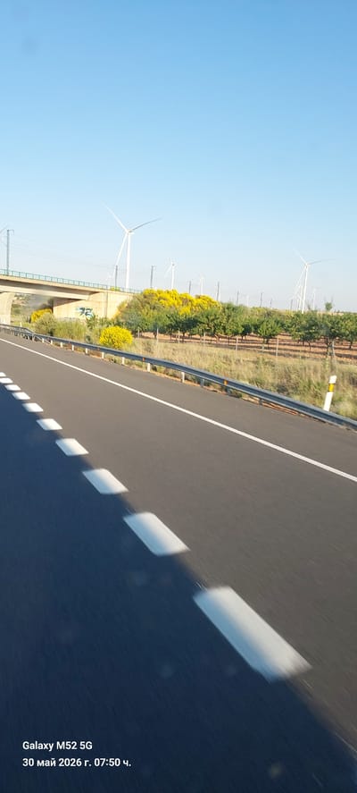 A photograph taken from a moving vehicle, possibly a car, shows a view of a highway and a landscape with wind turbines. The location is likely near Motilla del Palancar, Spain, suggested by the landscape and infrastructure. The highway asphalt appears smooth and new, with dashed white lane markings dividing the road into lanes. A metal guardrail runs along the side of the road, separating the highway from the adjacent terrain. Beyond the guardrail, there is vegetation consisting of trees and bushes, some with yellow flowers. Further in the distance, wind turbines are visible, indicating the presence of a wind farm. The sky is clear and blue. The photograph seems to have been taken on the 30th of May in 2026 at 07:50, as indicated by the phone's timestamp in the bottom right corner.