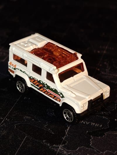 A white toy off-road vehicle with orange and green decals is placed on a black surface with a faint map-like pattern. The toy vehicle has a detailed interior and a rack on the roof containing amber-colored, translucent objects that appear to be logs. The off-road decals on the side of the vehicle include the text "4x4" and "OFF ROAD". The surface beneath the toy has faint lines and markings, and the word "FINLAND" is partially visible in white lettering, suggesting it is a map. The lighting is dim, creating shadows and highlighting the toy. The scene does not depict any people or actions. The location in Dörpen, Germany is not specified or indicated within the image itself.