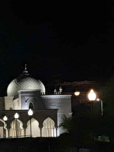 The image depicts a mosque at night in Privolzhsky, Russia. The mosque features a prominent dome with an intricate lattice pattern and a golden finial. The building is illuminated by external lights, highlighting its white facade and arched colonnades. Several street lamps with glowing tops are visible in the foreground, casting a warm light. The sky above is dark, with some faint clouds illuminated by distant light sources, possibly the moon. The scene is otherwise dark, with the surrounding vegetation appearing as silhouettes. There are no people visible in the image.