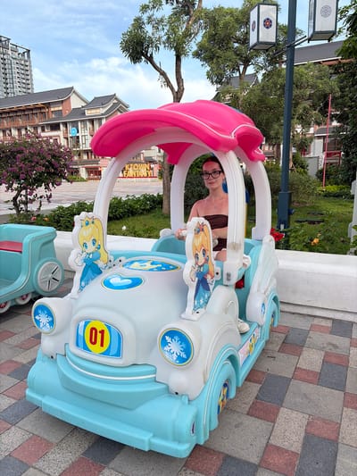 A young woman wearing glasses and a brown strapless top sits in the driver's seat of a light blue and white toy car with a pink canopy. The car is decorated with cartoon princess figures, snowflakes, and a yellow oval with the number "01" inside. She is holding the steering wheel, and her body is positioned as if she is driving. The car is on a paved area with a patterned stone surface. In the background, there are buildings with traditional Asian-inspired architecture, trees, and a tall modern building. The sky is partly cloudy. The environment appears to be a public outdoor space, possibly a park or a recreational area.