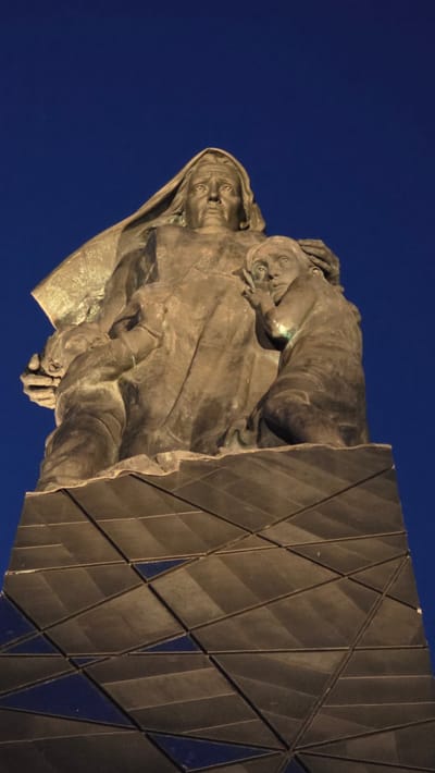 A low-angle shot shows a monument or sculpture, positioned against a solid blue sky. The sculpture depicts a woman holding a child, rendered in a weathered bronze or similar material. To the right of the sculpture is a large, geometric structure, composed of numerous triangular and diamond-shaped panels. The monument is located in Izhora (Verevskoe S/P), Russia.