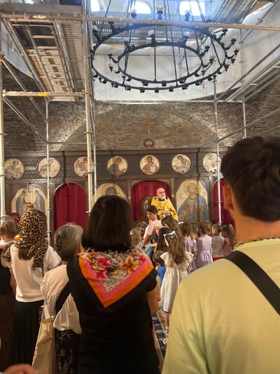 The image depicts the interior of a church, specifically the Church of Saint-Charles Borromeo in Montgeron, France, undergoing renovation, as evidenced by the scaffolding present. The scene is viewed from behind a crowd of people, mostly women and children, facing towards the altar area. A clergyman, likely a priest, dressed in a golden robe and a white vestment, stands on a raised platform, facing the congregation. He is positioned in front of a large iconostasis, which features multiple religious icons in circular and rectangular frames. The icons depict religious figures, possibly saints or biblical characters. In the background, an ornate chandelier is visible, also partially obscured by scaffolding. The church's architecture features exposed brickwork and arched ceilings. The congregation appears to be observing a religious service or ceremony.