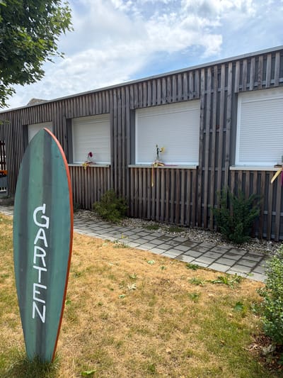 In Ufhusen, Switzerland, an outdoor scene captures a building with a modern, vertical wooden slat facade and three white roller shutters. In the foreground, a tall, oval-shaped, green and red sign with white lettering reading "GARTEN" stands upright. To the right of the sign, a paved pathway made of square gray stones leads towards the building, bordered by dry, yellowed grass and small green bushes. Yellow ribbons and a small vase with a flower are visible on the windowsill of the middle window. The sky above is partly cloudy with white cumulus clouds.