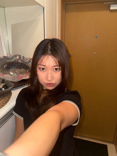 A young woman poses in a selfie, tilting her head. She has long, dark hair and wears a dark shirt with white trim on the collar and sleeves. In the background, there's a plain beige door and what looks like a closet or doorway leading to a darker space. Various items are visible in the foreground including a colorful hat. The overall lighting appears to be artificial.
