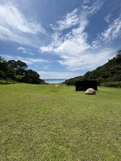 The image shows a wide, green lawn in front of a body of water. In the foreground, there is a large, rounded boulder placed in front of a black, rectangular structure that appears to be an art installation. The lawn slopes gently towards the water, which is calm and reflects the blue sky with scattered white clouds. Behind the lawn, a dense forest covers a hillside that meets the water. In the distance, across the water, there are faint outlines of mountains. The sky is a clear blue with wispy clouds. The scene is likely part of the art installations on Naoshima Island, known for its contemporary art museums and outdoor sculptures.