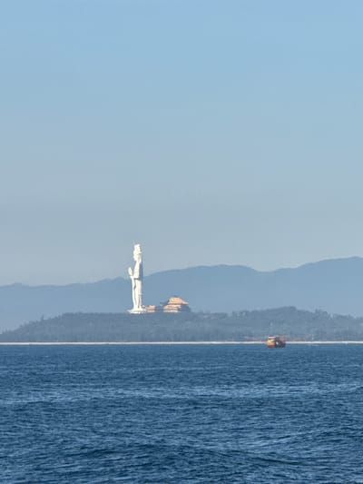 The image shows a wide, blue ocean in the foreground. In the middle ground, a verdant, tree-covered hill rises from the sea. Atop the hill stands a tall, white statue of a figure with a headdress and hands clasped. To the right of the statue, a traditional East Asian-style building with a gold-colored roof is visible. In the distance, a range of hazy blue mountains forms the backdrop. A small, colorful fishing boat with red and yellow accents is visible on the ocean, to the right of the hill. The sky is a clear, pale blue. This scene is likely the Lady Buddha statue at Son Tra Peninsula in Da Nang, Vietnam, near An Hải.