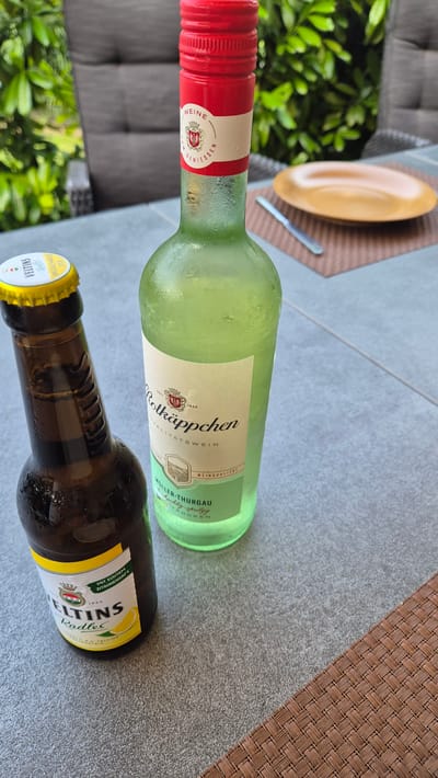 This is a close-up, slightly angled photograph of two bottles on a grey, tiled surface, likely an outdoor table. The bottle in the foreground, on the right, is a brown glass beer bottle with a yellow and white label. The text on the label includes "VELTINS" and "Radler," indicating it's a radler (a beer mixed with lemonade or fruit soda). There is also text that reads "MEGACHANCE 2025" and "MIT ECHTEM ZITRONENSAFT" (with real lemon juice). The bottle appears to be cold, with condensation visible.

Behind the beer bottle and slightly to the left is a green glass bottle, likely wine, also covered in condensation. It has a light-colored label with red and black text. The visible text includes "Rotkäppchen," "WEINQUALITAT," "MÜLLER-THURGAU," and "ROCKEN" (dry). The red cap of this bottle is prominent.

In the background, out of focus, are green foliage, a gray cushioned chair or sofa, and a brown woven placemat with a yellow plate on it. The lighting suggests it might be daytime, possibly outdoors, given the greenery and the cushioned furniture. The overall scene implies a relaxed setting, perhaps a meal or break outdoors. The location is identified as Dissen am Teutoburger Wald, Germany.