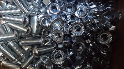 The image displays a close-up, overhead view of a large pile of metallic fasteners, predominantly bolts and nuts, scattered in a bin or container. The fasteners are shiny and appear to be made of steel or a similar metal, reflecting the light. The bolts are silver-colored with visible threads and some have hexagonal heads, while others have rounded heads with slotted drives. The nuts are mostly hexagonal with flanged bases that have a serrated or ridged texture, designed to prevent loosening. The fasteners are densely packed, creating a textured surface with many overlapping components. Some fasteners are partially obscured by others, and the lighting creates strong highlights and shadows, emphasizing the three-dimensional nature of the pile. There are no people visible in the image, and the context suggests a storage or supply situation for hardware. The image does not provide any information about the location in the city of Moulins-sur-Orne, France, as no recognizable landmarks or geographical indicators are present.