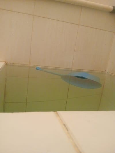 The image shows a light blue ladle submerged in yellowish-green water in a bathtub. The bathtub is tiled with light beige square tiles. A metal bar is visible at the top of the bathtub. The scene appears to be in a bathroom. There is no indication of the location being in Surabaya, East Java.
