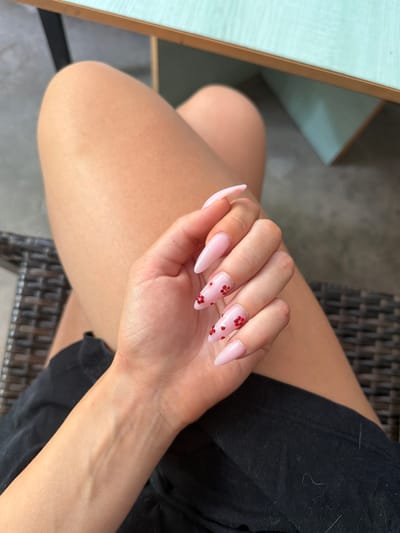 The image displays a close-up of a person's hand and leg. The hand is positioned over the person's thigh, which is covered by black fabric. The nails on the hand are long and almond-shaped, painted a light pink color, and adorned with small red rhinestones. The background suggests an outdoor setting, possibly a patio or seating area, with a table partially visible at the top. The location is stated to be Prosena, Bulgaria.