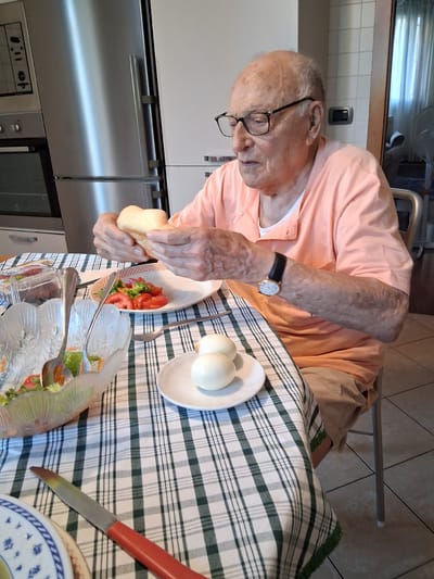 A man is sitting at a table with a green and white checkered tablecloth, preparing to eat. He is wearing glasses and an orange shirt. On the table, there is a plate with tomatoes and lettuce, a small dish with two hard-boiled eggs, and a bowl of salad. The man is holding a slice of bread in his hands. A knife with a red handle is lying on the table. The scene appears to be indoors, possibly a kitchen or dining room, with a stainless steel appliance visible in the background. The lighting suggests it could be daytime. The man's focus is on the food, and his expression is neutral. The location is identified as Padua, Italy.