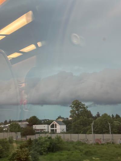 The image appears to be taken from inside a vehicle, possibly a train, looking out a window. Reflections on the window suggest interior lighting, with rectangular, illuminated panels visible in the upper left portion. The exterior scene depicts a suburban or semi-rural environment under a cloudy, possibly stormy, sky.
In the mid-ground, there are several houses, one of which is a modern, white structure with a distinctive A-frame roof. Trees are scattered throughout the landscape, with denser foliage forming a line along the mid-ground, behind a fence. The fence appears to be a noise barrier, common along transportation routes.
In the foreground, there is a grassy area with more greenery. The overall atmosphere is somewhat somber due to the heavy cloud cover. There are no visible people or specific actions taking place. The location is identified as Luboń, Poland.