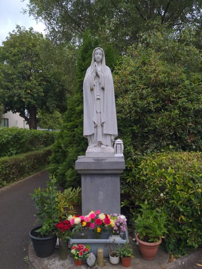 The image depicts a stone statue of the Virgin Mary, likely located in an outdoor setting in Ballymena, United Kingdom. The statue stands on a pedestal with a cross and a circular emblem. In front of the pedestal, there are several potted plants and vases of flowers, including red roses and a mix of pink, yellow, and white flowers. A small white lantern sits on the side of the pedestal. Behind the statue, there are lush green bushes and trees. A paved path runs along the left side of the image, and a building can be seen in the background. The overall scene suggests a memorial or a religious shrine in a garden or park.
