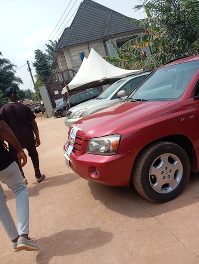 The scene depicts an outdoor environment in Okpatu-Ukehe, Nigeria, under clear, bright daylight. The foreground consists of reddish-brown unpaved dirt ground, marked by shadows cast from the left. A red Toyota Highlander SUV occupies the right foreground, parked facing right, with white and blue decorative sashes draped across its front grille. A light grey SUV is parked behind it, followed by a dark-colored sedan partially obscured by a white, pyramid-shaped canopy tent.

In the mid-ground, a person wearing a dark brown tunic and a dark cap is visible from behind, walking towards the right. Another person's lower body is visible in the extreme left foreground, wearing light blue jeans and grey slip-on shoes, also facing right.

The background features a multi-story building with light-colored walls and a dark grey tiled roof, partially visible behind a decorative concrete gate. A wooden utility pole with overhead lines stands on the left. Green foliage, including palm trees and large-leafed plants, is present throughout the background.