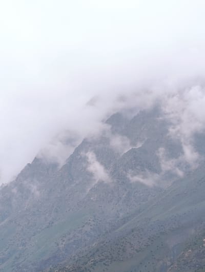 The image shows a mountain landscape in Darcha, India. The mountains are covered in mist and clouds, obscuring the peaks and creating a dramatic atmosphere. The slopes of the mountains are covered with sparse vegetation, with some rocky outcrops visible. The sky is overcast and appears to be a pale white, blending into the mist. The overall impression is one of a remote and rugged environment, shrouded in atmospheric conditions.