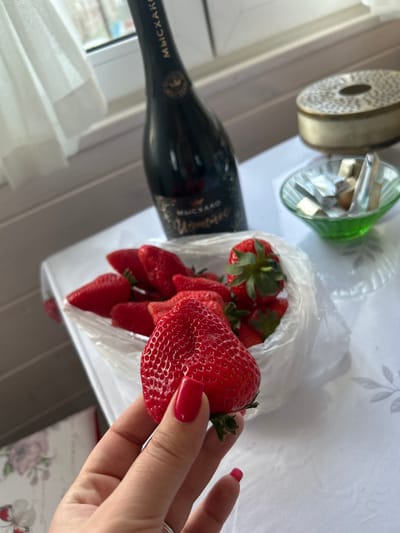 The image shows a still life arrangement on what appears to be a table indoors, potentially near a window. There is a bottle of sparkling wine, possibly champagne, with Cyrillic script visible on the label, identifying it as "Abrau-Durso," which is a Russian brand. This would indicate a possible location in Russia, though the image does not specify a location in Bashur.

Next to the bottle is a clear glass object, possibly a decorative vase. A clear plastic bag filled with fresh strawberries is centrally positioned. A hand with red nail polish holds a large strawberry. A roll of ribbon is visible in the background. The overall arrangement suggests a celebratory or festive occasion.