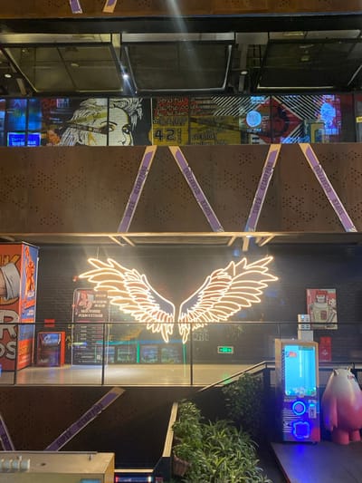 This image captures an indoor scene, likely within a modern entertainment venue, arcade, or themed commercial space, illuminated by artificial light, suggesting it's nighttime or evening. The perspective is slightly angled, looking across a room with various decorative and functional elements.
Dominating the center of the frame is a large, brightly glowing neon sign in the shape of stylized wings, emitting a warm, light yellow or white light against a dark background. To the left of these neon wings, a series of dark brown, perforated panels act as a partition or wall. Diagonally stretched across these panels are multiple purple tapes or banners, each bearing the repeated text "SUPER 42 CARP POND."
Further to the left, behind these panels, is a wall adorned with an eclectic mix of pop-art style murals and posters. These feature comic-book-like illustrations, bold graphics, and fragmented text. Visible phrases and words include "pendful," "Call the powe," "Rus has been desti," "CARP," "42," "EVERYDAY," and "Gas Dam 10 AM," along with "COLE," "SC," "D," "W," and repetitions of "SUPER 42 CARP POND." One distinct illustration shows a portion of a face, possibly a woman's.
To the right of the neon wings, the scene shifts to an area featuring what appear to be interactive gaming stations or arcade machines. One prominent machine has a bright, colorful screen displaying graphics, accompanied by an array of buttons and controls. Above this machine, a large, smooth, light pink, stylized pig figurine or sculpture is positioned. Further right, a planter filled with lush green foliage adds a touch of natural element to the otherwise urban and neon-lit environment.
Reflections are visible on what appears to be a large glass partition or window in the central part of the image, reflecting some of the elements from the opposite side of the room, including the bright screens of arcade machines and other lighting. Text seen on or reflected from this area includes Chinese characters "不限" (Bù xiàn - Unlimited/No limit) and "先到K打台球" (Xiān dào K dǎ tái qiú - First come, first served for K / playing billiards), suggesting activities offered at the establishment. Other fragmented text includes "论" (Lùn - theory) and "質" (Zhì - quality), and a stylized "ΓΟΛΕΟ".
The overall atmosphere is vibrant and contemporary, characterized by the interplay of dark surfaces, bright neon, and colorful digital displays, creating a setting for entertainment and leisure.