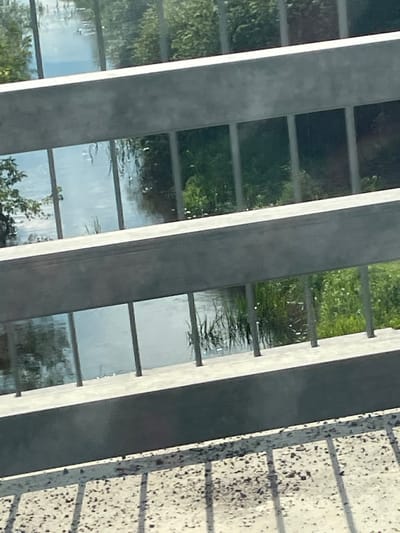 The image shows a view through a metal railing, likely on a bridge or walkway, looking down at a body of water. The water reflects the sky, trees, and greenery. The railing consists of horizontal bars and vertical supports. The ground in the foreground is textured, possibly concrete or pavement, with shadows cast by the railing. The scene is set outdoors, in a natural environment with water and vegetation. The location in Yaltsivka, Ukraine, suggests a rural or semi-rural setting. There are no people or specific actions depicted in the image.