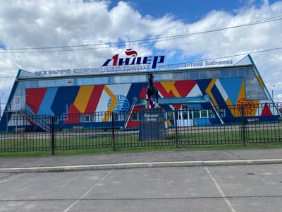 The image depicts the "Лидер" (Leader) Sports and Recreation Complex named after K. Baymeev, located in Ust-Ordynsky, Russia. The building features a colorful facade of blue, red, yellow, and white geometric shapes. Above the entrance, signage indicates the complex's name in Russian. A black horse statue stands behind a black metal fence in front of the building, with a plaque reading "Степной ветер" (Steppe Wind). The ground in front of the fence is paved with grey concrete, and some greenery is visible behind the fence. The sky above is partly cloudy.