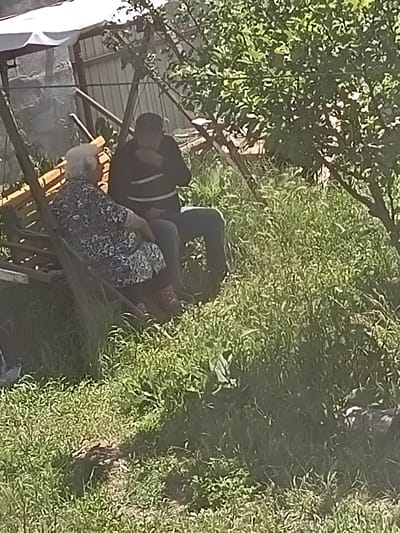 In Nor Geghi, Armenia, an elderly woman with white hair, wearing a floral print dress, sits on a wooden swing. Beside her, a man wearing a dark long-sleeved shirt with horizontal stripes sits on the same swing, his head bowed as if eating or speaking. They are surrounded by lush green grass and foliage, with a tree's branches casting dappled shadows on the scene. A portion of a white canopy is visible overhead, and a fence can be seen in the background. The overall setting suggests a relaxed outdoor gathering in a rural or semi-rural environment.