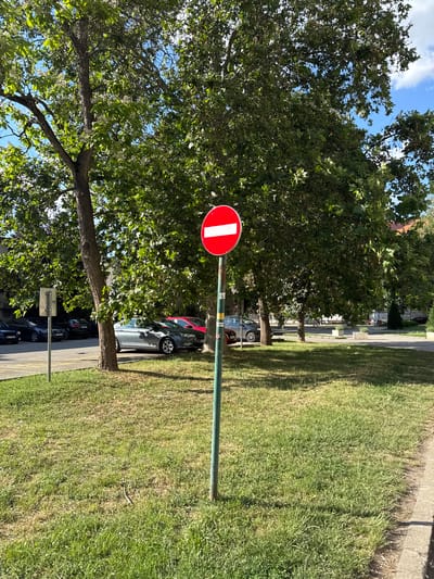 The image displays an outdoor scene in Becej Municipality, Serbia, captured under bright daylight conditions with a blue sky containing scattered white cumulus clouds. In the immediate foreground, a green grassy area exhibits varying shades of green and patches of lighter, drier grass. A prominent "No Entry" traffic sign, characterized by a red circle with a central horizontal white bar, is mounted on a faded green pole and positioned slightly to the right of the frame's center, casting a shadow to its right. A concrete curb lines the bottom right edge of the frame.

The midground is dominated by multiple large deciduous trees featuring dense green foliage, providing substantial shade. One tree on the left exhibits lighter green leaves and white blossoms, while other prominent trees display broad, lobed leaves. Behind the grassy area and trees, a paved asphalt surface is visible, functioning as a road or parking area. Several vehicles are parked: a dark blue/black sedan (an Audi) is visible on the far left, a silver-grey sedan (a Ford Focus) is partially obscured by a tree trunk slightly right of center, and a red car is discernible further in the background. A secondary metallic signpost with an indecipherable white rectangular sign is located in the grassy area to the left.

The background further reveals a paved area that appears to be a pedestrian pathway or a section of a park, featuring several light-colored concrete planters and benches. Indistinct human figures are visible seated on these benches in the distance. Architectural structures, characterized by red-tiled roofs, are partially visible through the tree canopy in the far background. The scene is illuminated by strong sunlight, producing clear, defined shadows from trees, the sign pole, and other objects.