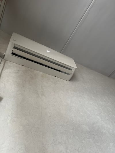 The image shows a white air conditioning unit mounted on a textured, light-colored wall. The unit is positioned at an angle, with its louvers partially open. A faint Apple logo is visible on the front of the unit, suggesting it might be a specialized device or have some Apple integration. The wall has a rough, uneven texture, and there are some faint lines or cracks visible on the ceiling in the upper left portion of the image, which is painted a lighter shade of gray. The lighting in the image appears to be artificial, casting subtle shadows. No people are visible in the frame. The overall scene suggests an indoor environment, possibly a room in a residential or commercial building. A small sticker with text is visible on the lower right edge of the air conditioning unit, but the text is too small to be legible. The color palette is dominated by shades of white and gray.