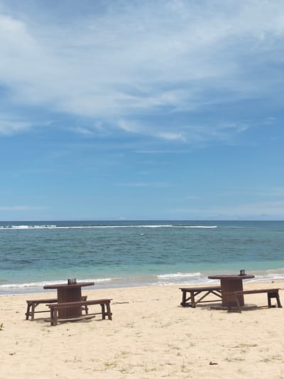 The image shows a beach scene in Monikeun, Indonesia. Two wooden picnic tables are situated on the sandy shore. The ocean is visible in the background, with small waves approaching the beach. The sky is blue with scattered clouds.