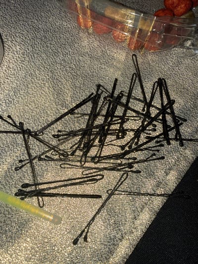A cluttered arrangement of black bobby pins is scattered on a textured, silver-grey fabric surface. Some pins overlap and intersect. A clear plastic container filled with reddish-brown objects sits to the left. A cylindrical, translucent object with a yellowish tint lies among the bobby pins on the lower right. A dark area is visible in the upper right corner, likely indicating the edge of a surface or object.
