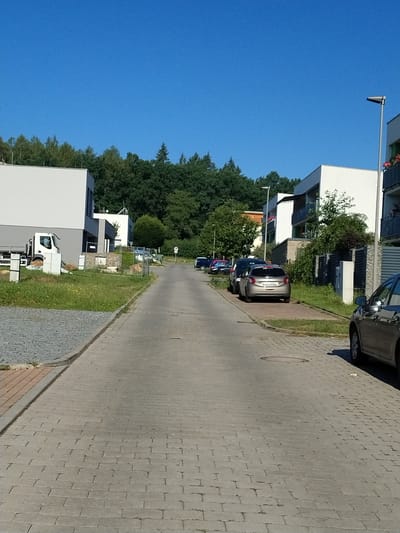 The image depicts a street scene in a residential area, likely in Písek, Czechia. The scene is oriented vertically, with the sky dominating the left side and the ground on the right. Modern buildings with white and grey facades, some with balconies, are visible on the left, interspersed with lush green trees. A paved sidewalk runs along the right side of the street, with a cobblestone pattern.

Several cars are parked along the street. One is a silver hatchback, and another is a darker colored sedan. In the foreground on the right, a white truck is partially visible. A street light is mounted on a pole extending from one of the buildings. The sky is a clear, bright blue, suggesting it is a sunny day. The time of day appears to be daytime, given the bright sunlight casting shadows.

Notable details include the architectural style of the buildings, which appear to be relatively new. The presence of trees and the well-maintained paving suggest a pleasant, orderly environment. No people are clearly visible in the image. No visible text is decipherable.